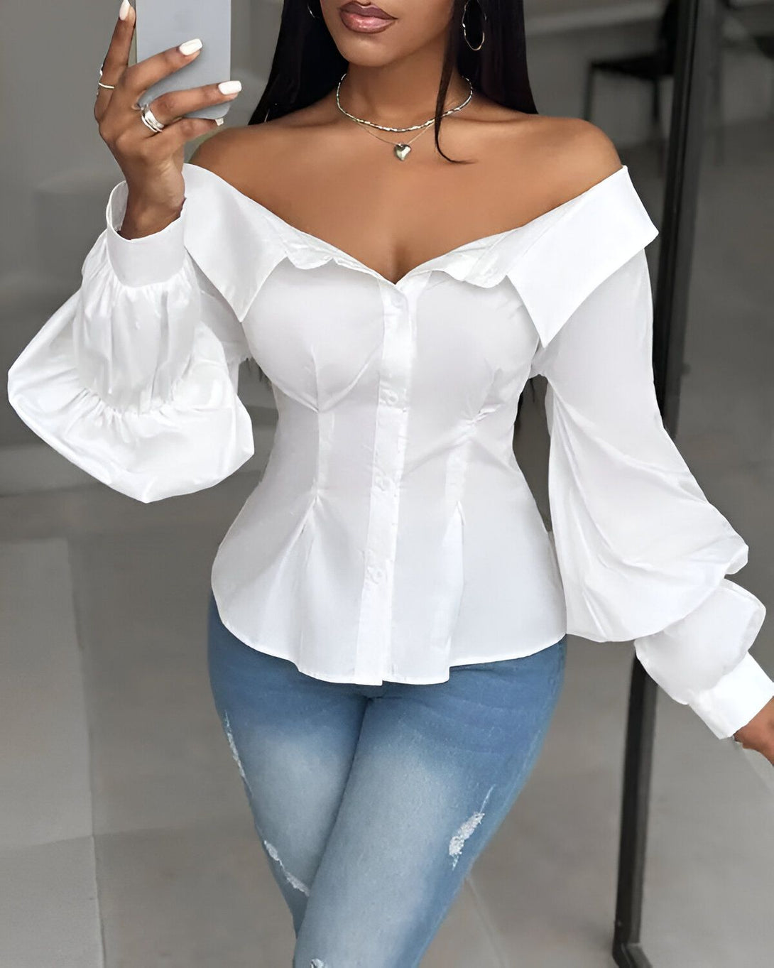 Une femme porte un chemisier blanc à épaules dénudées. Le chemisier est bouffant au niveau des manches et possède un col montant. Elle porte également un jean bleu déchiré.