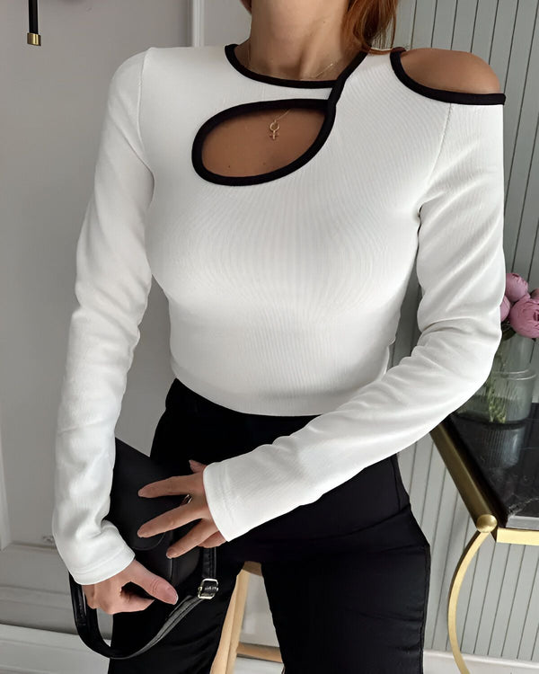 Une femme porte un haut blanc à manches longues avec une découpe sur l'épaule et un pantalon noir. Elle porte également un collier et un sac à main noirs.