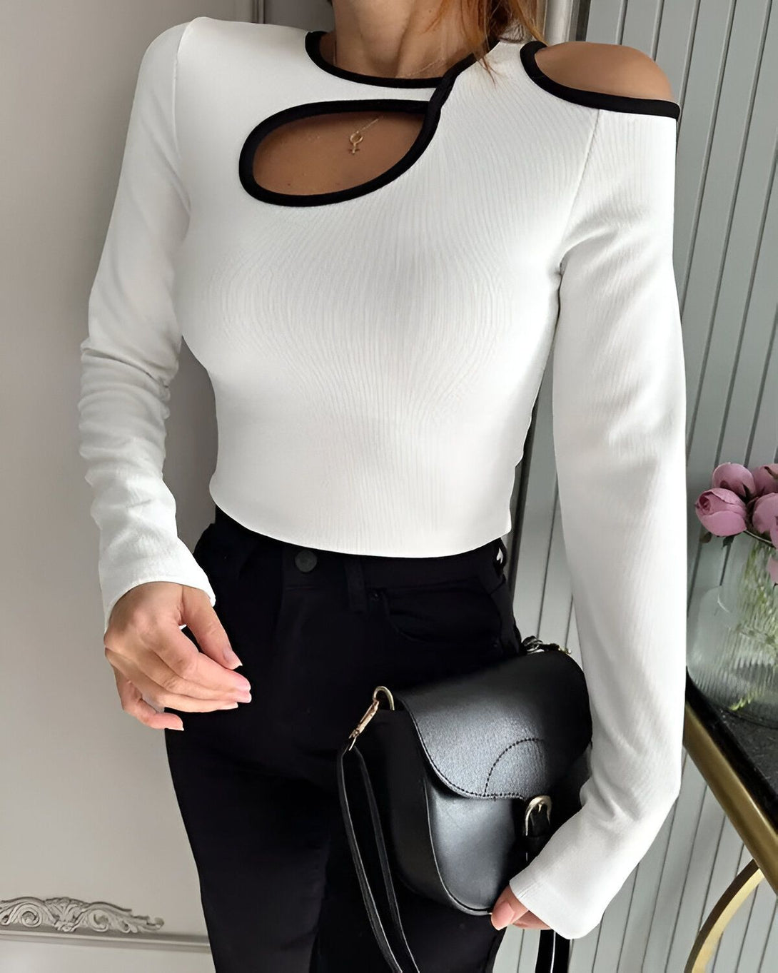 Une femme porte un haut blanc à manches longues avec une découpe sur l'épaule et un pantalon noir. Elle porte également un sac à main noir.