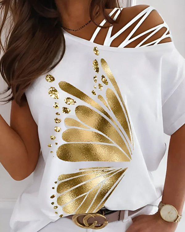 Une femme porte un t-shirt blanc avec un imprimé doré d'un papillon. Le papillon est sur le côté gauche du t-shirt et est fait de paillettes dorées. La femme a les cheveux longs et bruns et porte un collier noir. Elle est également porte une montre en métal argenté au poignet gauche.
