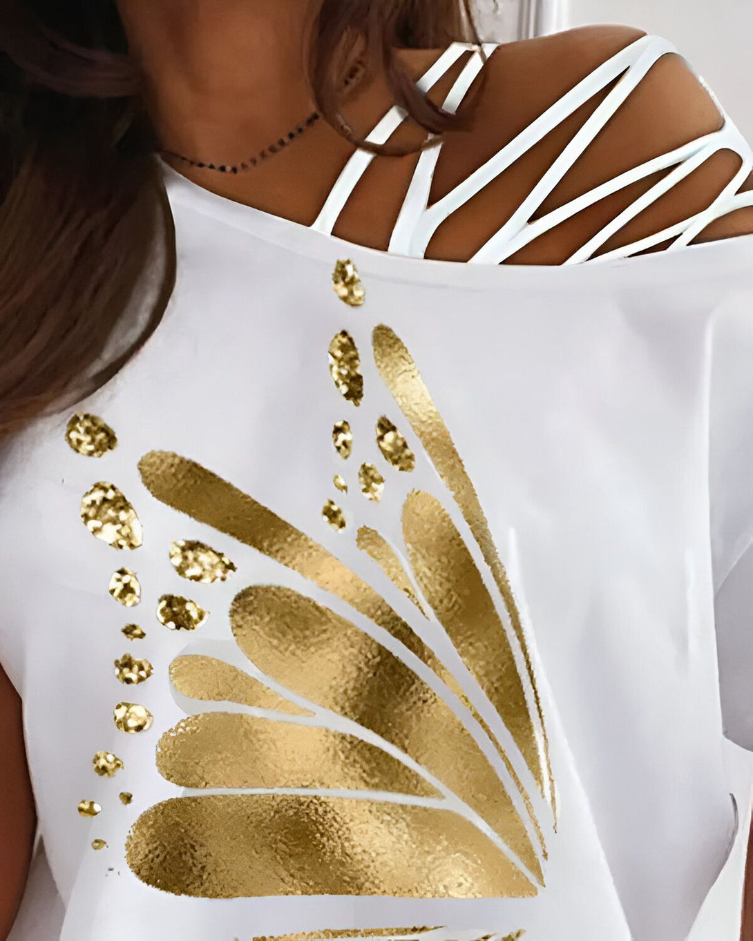 Une femme porte un t-shirt blanc avec un motif doré. Le t-shirt a une épaule dénudée et des lanières croisées sur l'autre épaule.