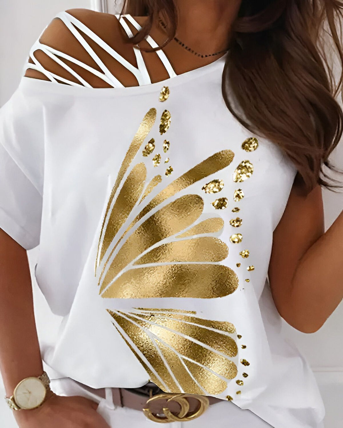 Une femme porte un t-shirt blanc avec un papillon doré imprimé dessus. Le t-shirt a des épaules dénudées et des lacets blancs qui se croisent sur le devant. La femme a les cheveux longs et bruns et porte un collier noir.