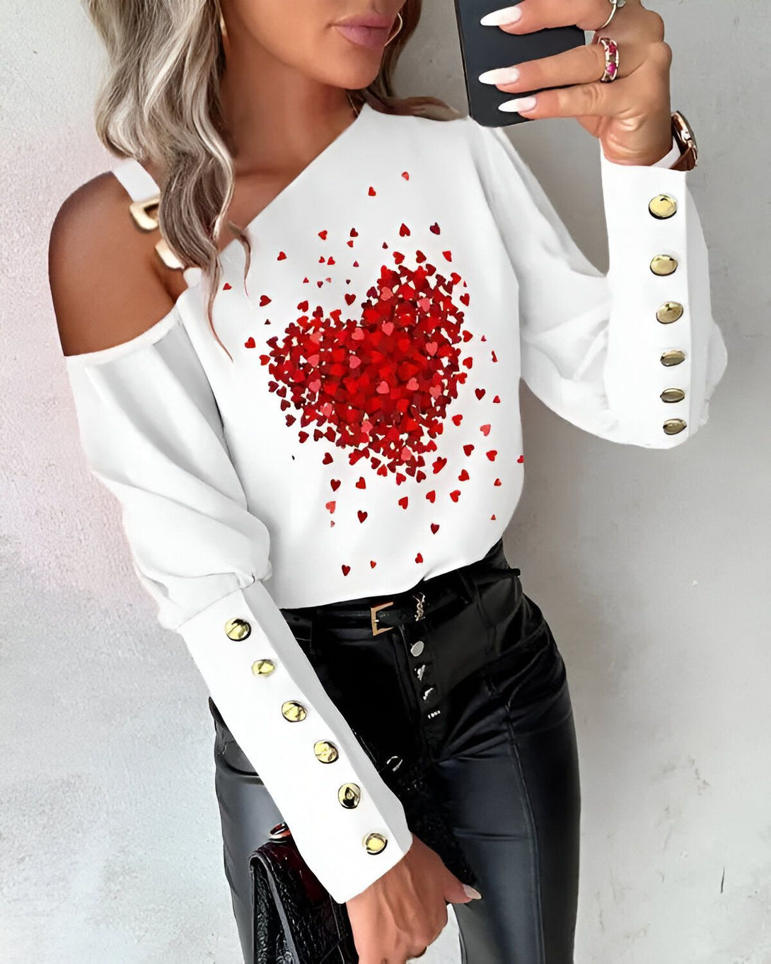 Une femme porte un chemisier blanc à épaules dénudées avec un imprimé de cœurs rouges. Le chemisier est rentré dans un pantalon noir. La femme porte également un collier et des boucles d'oreilles.