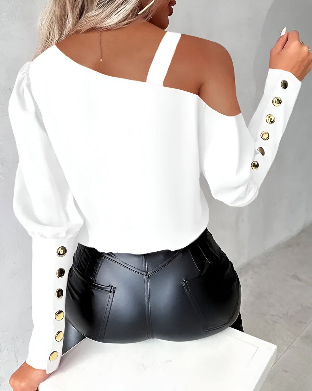 Une femme porte un chemisier blanc asymétrique avec des boutons dorés sur la manche gauche et une épaule dénudée. Elle porte également un pantalon en cuir noir.