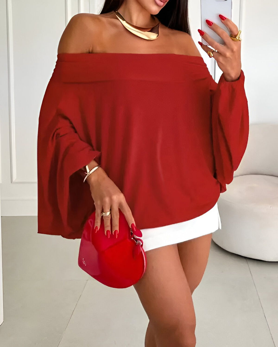 Une femme porte un haut rouge à épaules dénudées et un short blanc. Elle a un collier doré, des bracelets et une bague. Elle tient un sac à main rouge en forme de cœur.