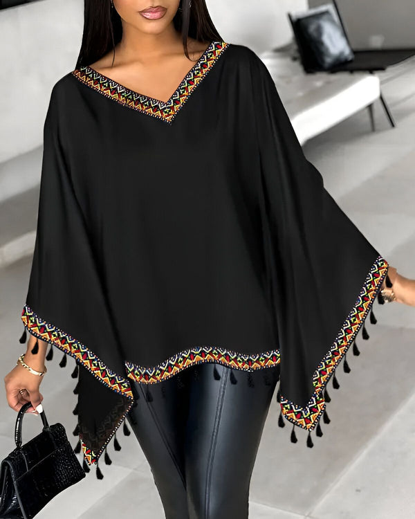 Une femme porte un chemisier noir avec des motifs colorés. Le chemisier est ample et a des manches chauve-souris. La femme porte également un pantalon en cuir noir et des talons hauts noirs.