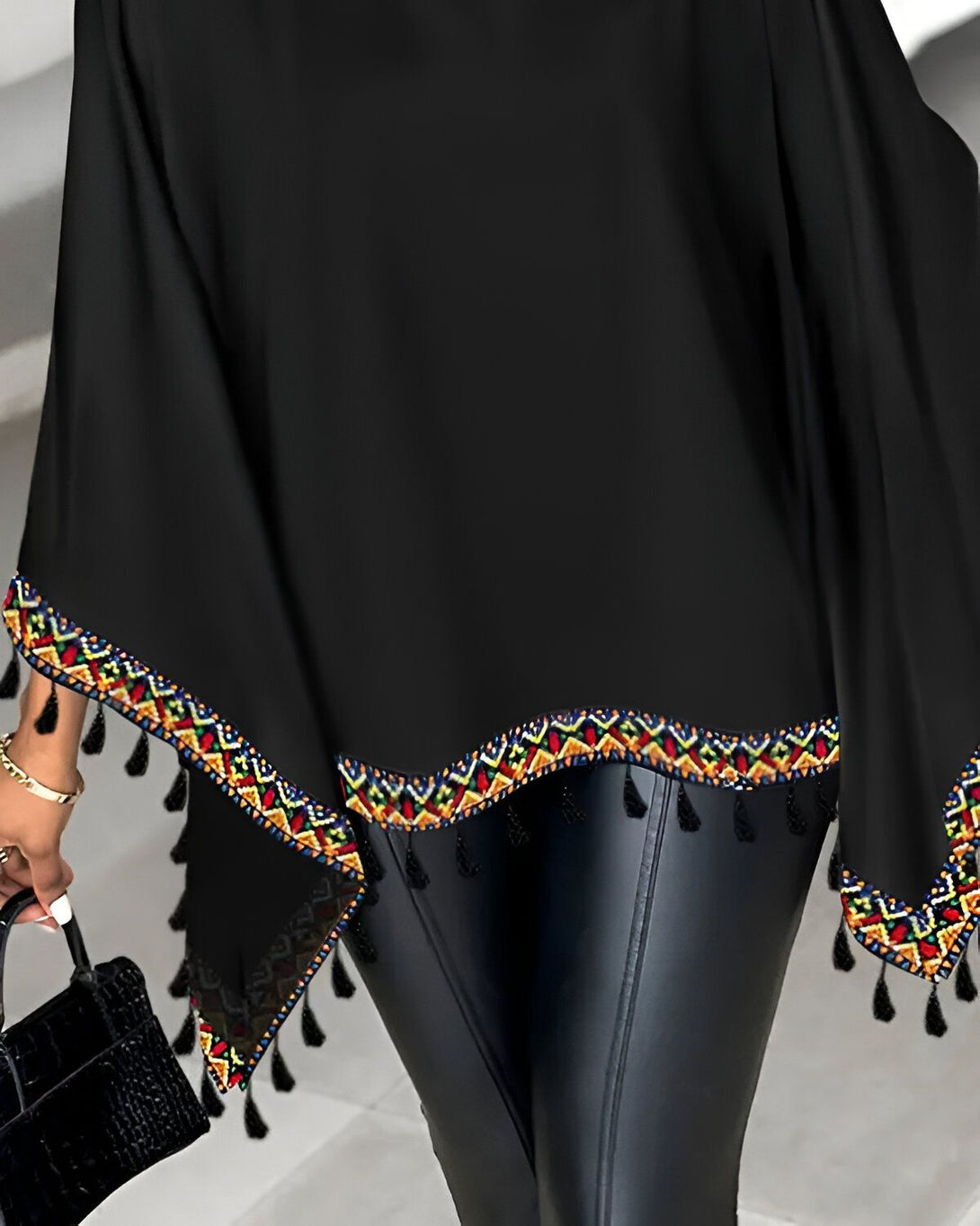Une femme porte un chemisier noir avec des broderies colorées et des franges au niveau de l'ourlet. Elle porte également un pantalon en cuir noir et des escarpins noirs.