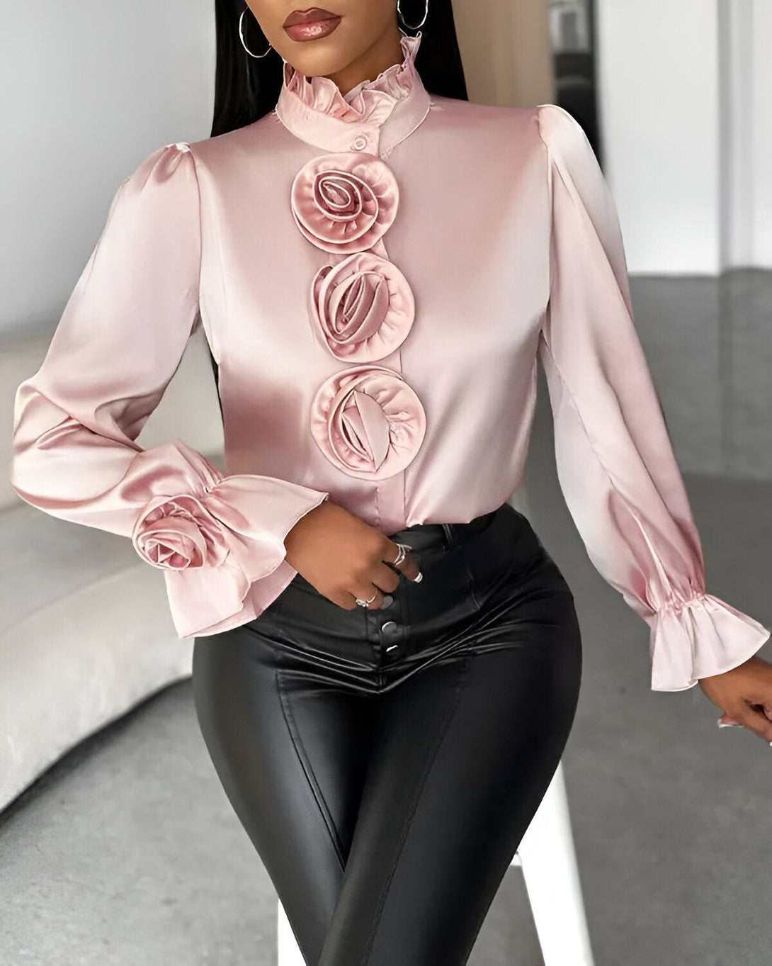 Une femme porte un chemisier rose en satin avec des boutons et des manches bouffantes. Le chemisier est orné de roses en satin de la même couleur. Elle porte également un pantalon en cuir noir et des talons noirs.