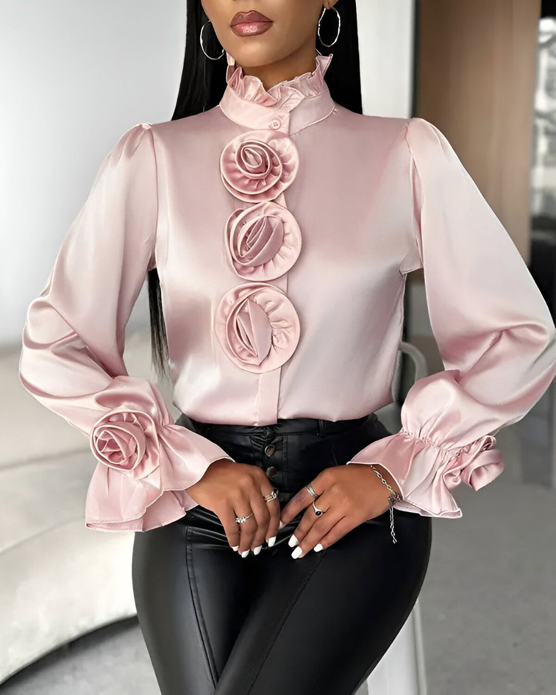Une femme porte un chemisier rose en satin avec des boutons en forme de roses. Le chemisier a un col montant et des manches longues bouffantes. Elle porte également un pantalon noir en cuir.
