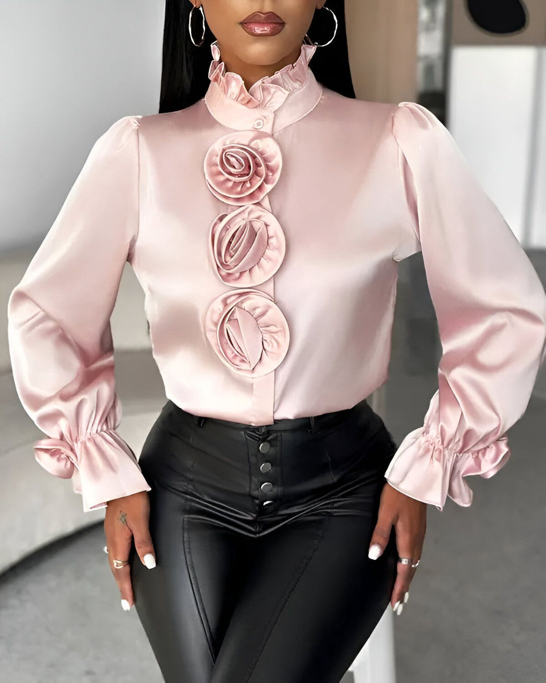 Une femme porte un chemisier rose en satin avec des boutons et des manches bouffantes. Le chemisier est orné de trois fleurs en satin de la même couleur. Elle porte également un pantalon noir en cuir.
