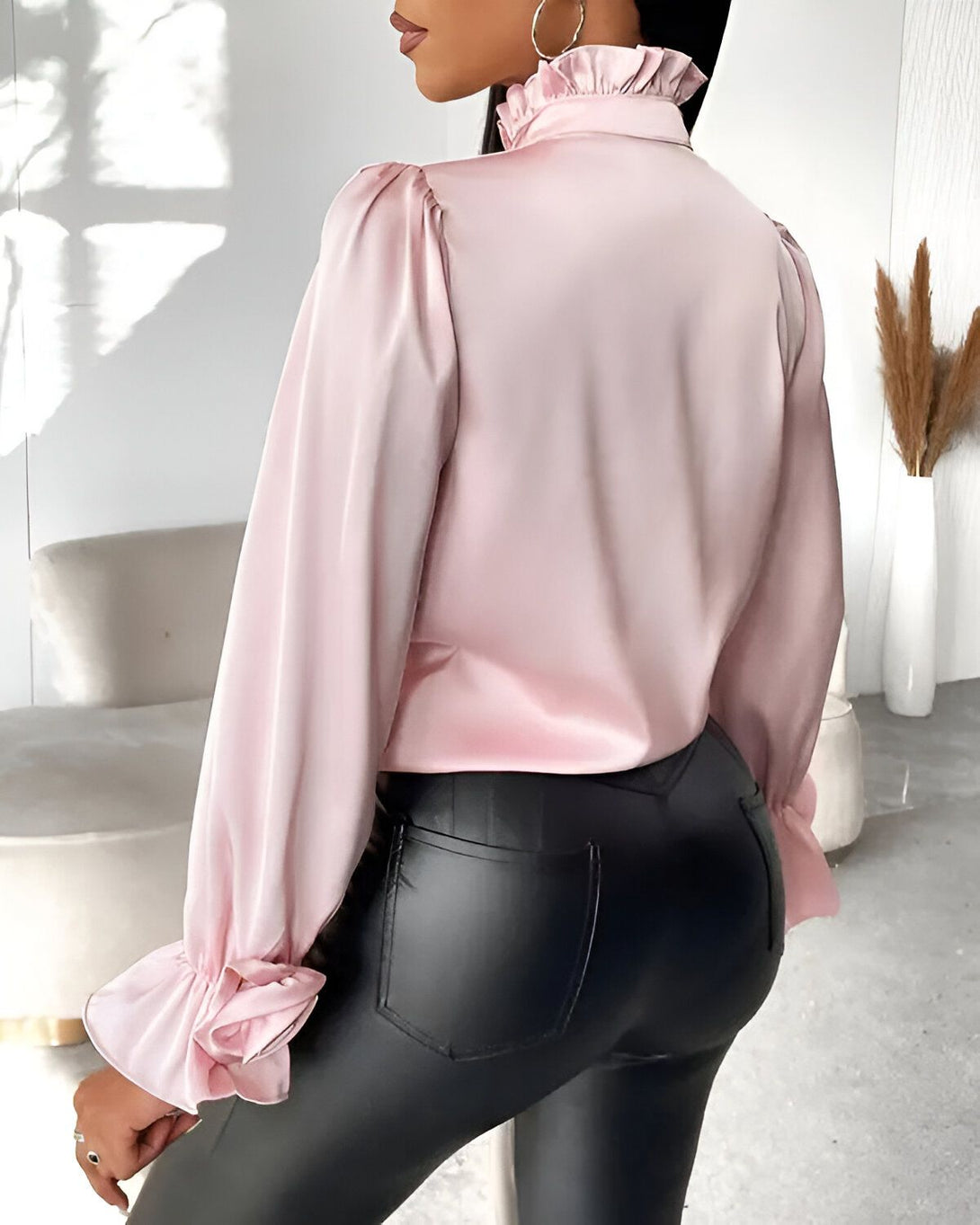 Une femme porte un chemisier en satin rose avec un col montant et des manches bouffantes. Elle est vue de dos et porte un pantalon en cuir noir.