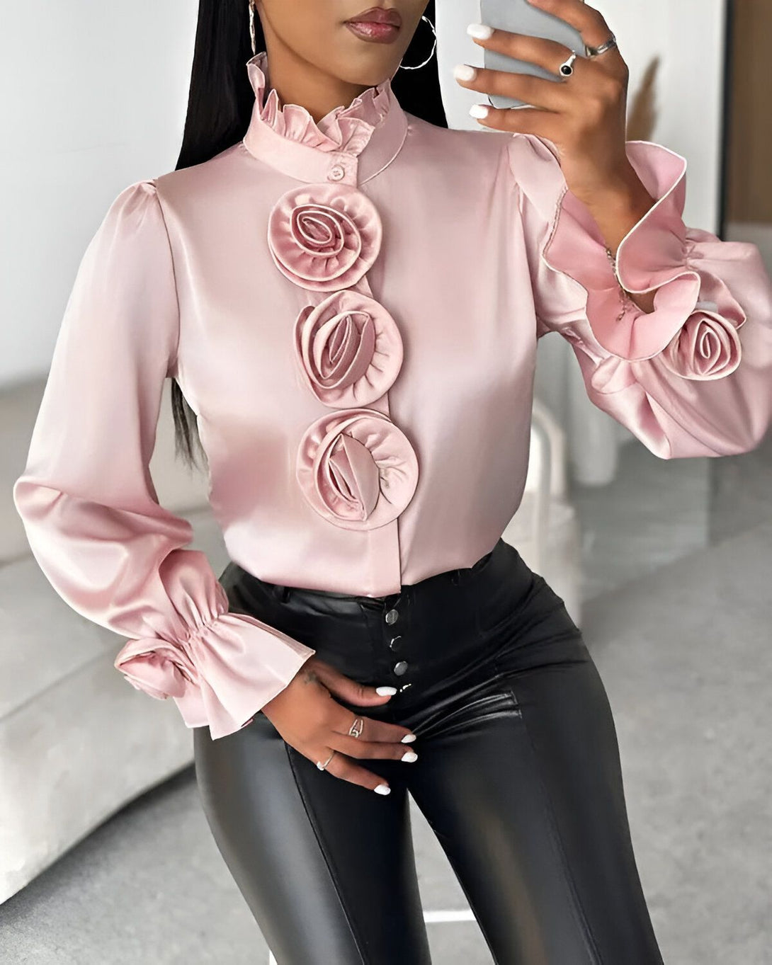 Une femme porte un chemisier rose en satin avec des boutons et des volants. Le chemisier est cintré à la taille et les manches sont bouffantes. Elle porte également un pantalon en cuir noir et des escarpins noirs.
