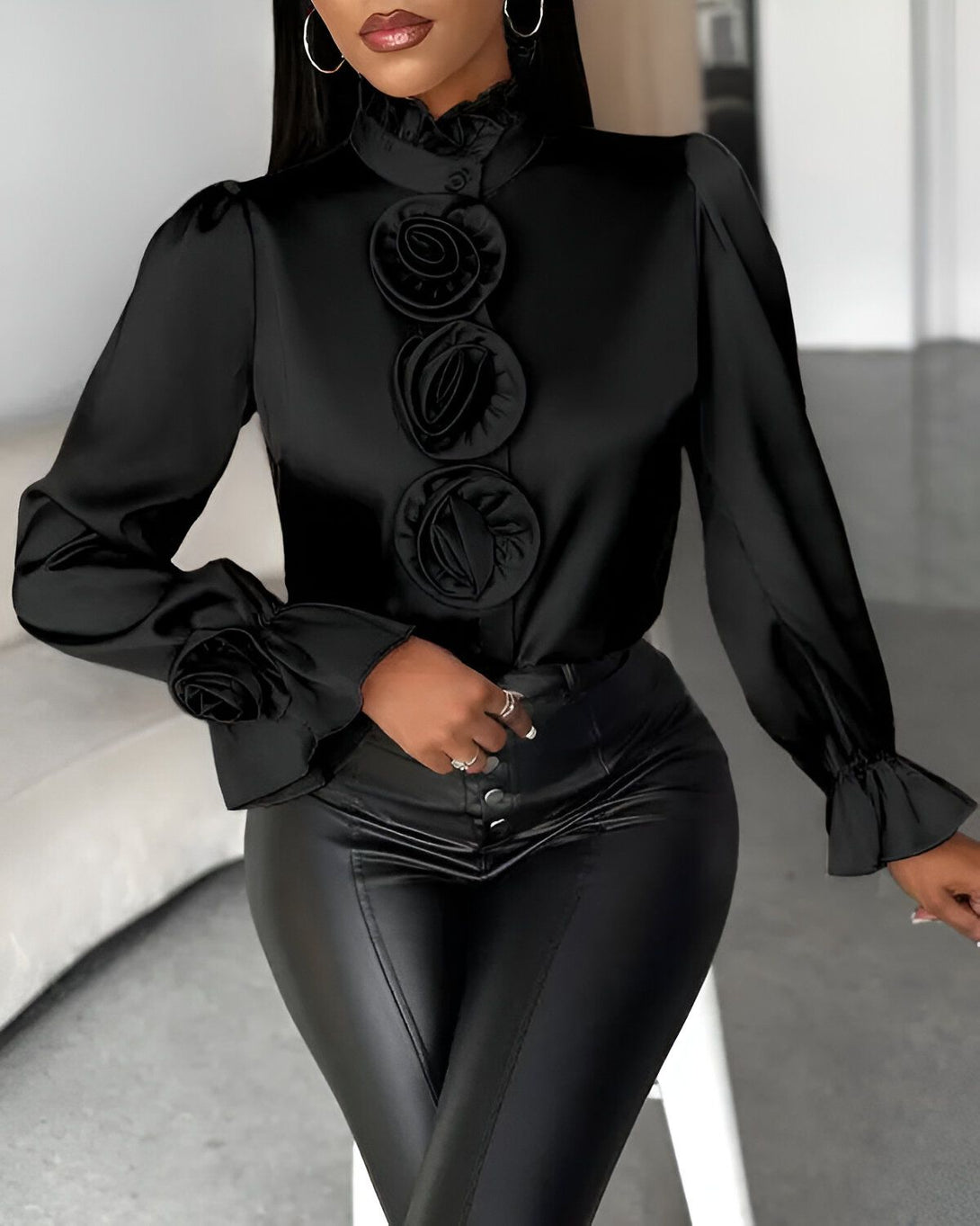 Une femme porte un chemisier noir en satin avec des boutons et des manches bouffantes. Le chemisier est orné de roses en satin noir. Elle porte également un pantalon en cuir noir.