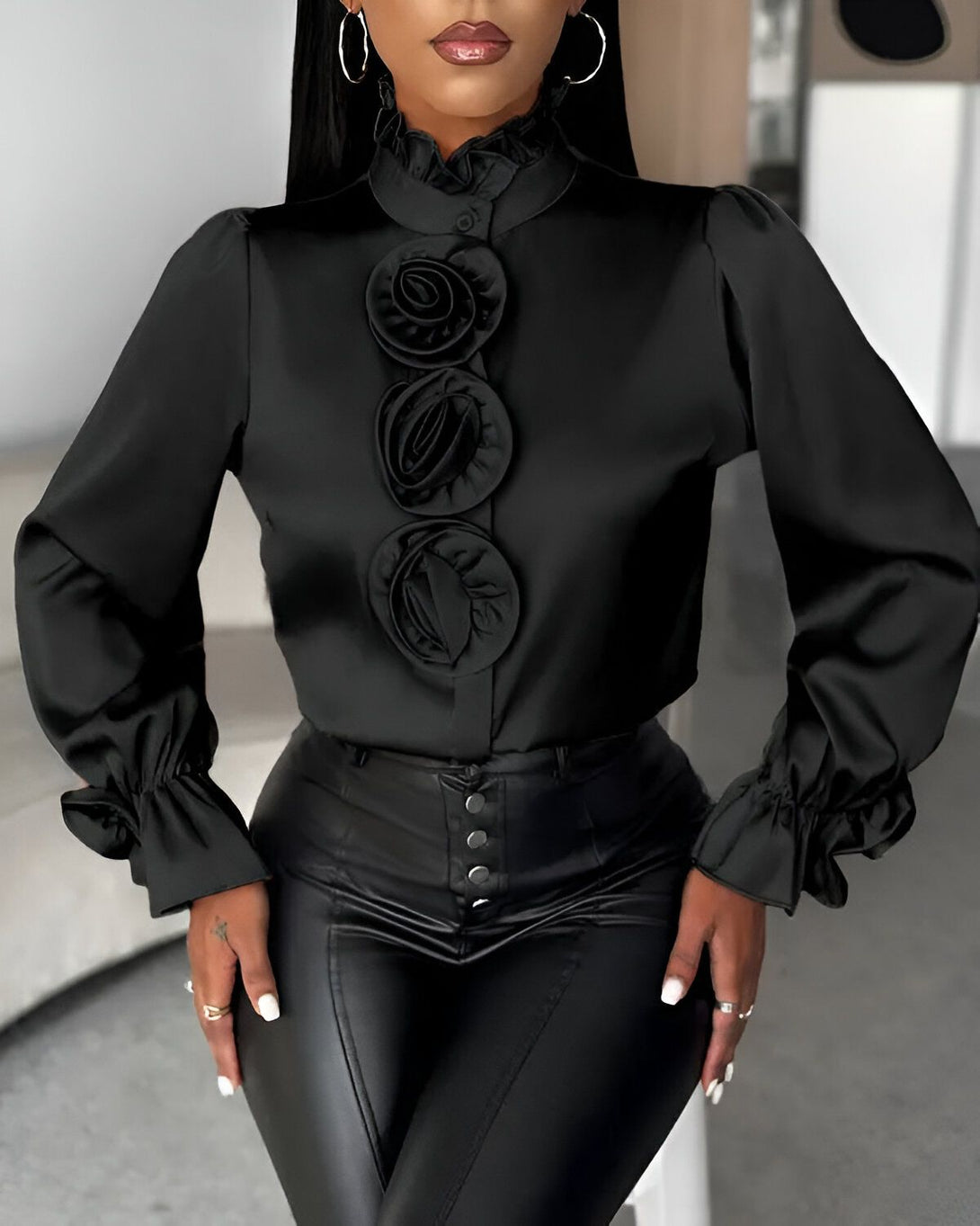 Une femme porte un chemisier noir en satin avec des boutons et des manches bouffantes. Le chemisier est orné de trois fleurs en satin noir. Elle porte également un pantalon en cuir noir.