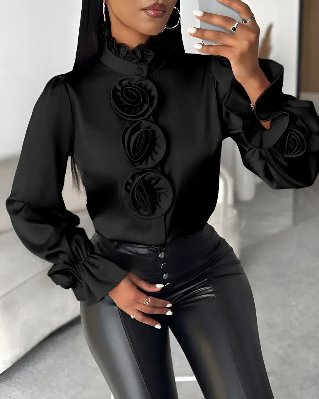 Une femme porte un chemisier noir avec des détails en forme de roses. Le chemisier est en satin et a un col montant. Les manches sont bouffantes et ont des poignets élastiqués. La femme porte également un pantalon en cuir noir et des talons noirs.