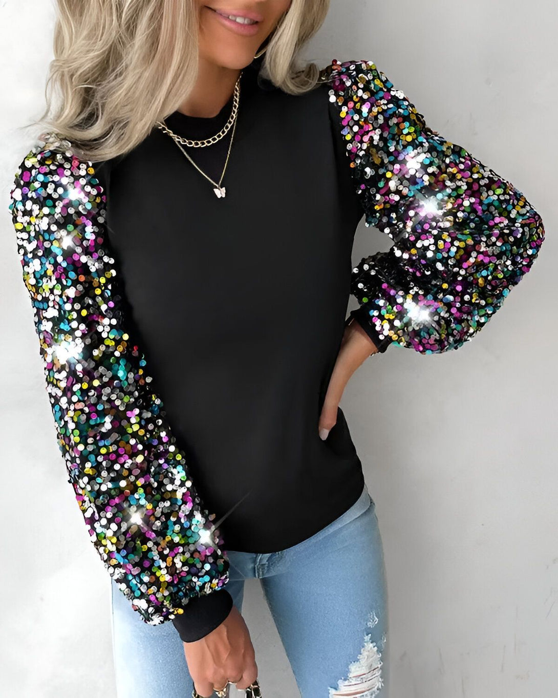 Une femme porte un haut noir à manches longues. Les manches sont recouvertes de paillettes multicolores. La femme est blonde et porte un collier avec un pendentif papillon. Elle porte également un jean bleu déchiré.