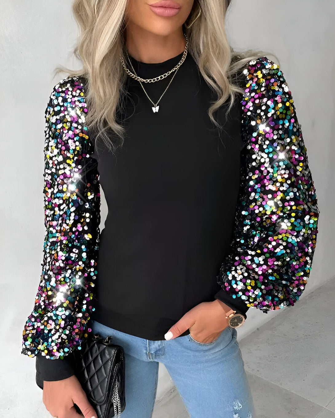 Une femme porte un haut noir à manches longues bouffantes et colorées. Les manches sont ornées de sequins multicolores. Elle porte également un jean bleu et un collier avec un pendentif papillon.