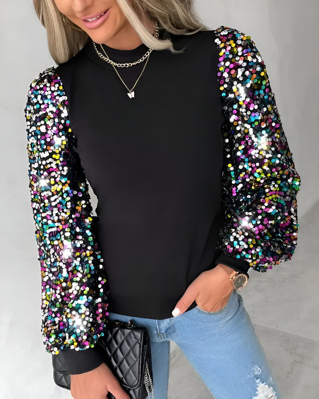 Une femme porte un haut noir à manches longues bouffantes et colorées. Le haut est rentré dans un jean bleu troué. Elle porte également un collier et une montre.