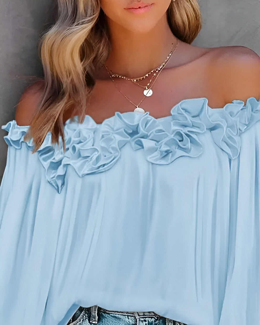 Une jeune femme porte un chemisier bleu clair à épaules dénudées. Le chemisier est en mousseline de soie et présente des volants sur les épaules. La femme a les cheveux blonds et porte un collier.