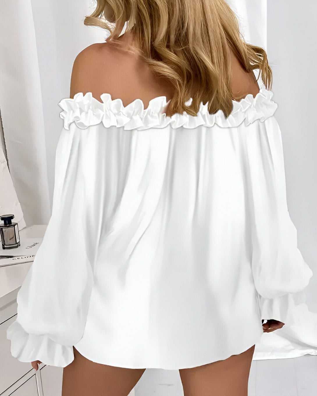 Une femme porte un chemisier blanc à épaules dénudées. Le chemisier est en mousseline de soie et a des manches bouffantes. La femme est blonde et a les cheveux longs. Elle est vue de dos.