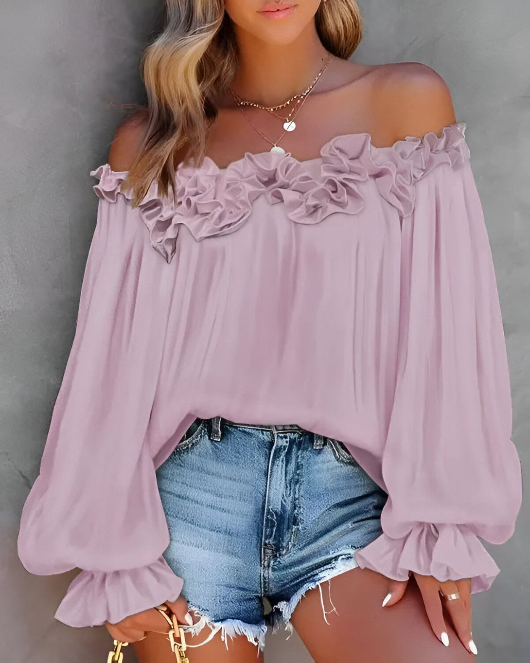 Une jeune femme porte un chemisier rose à volants et un short en jean. Le chemisier a des manches bouffantes et un col Bardot. La femme a les cheveux longs et blonds et porte un collier. Elle est debout devant un mur gris.