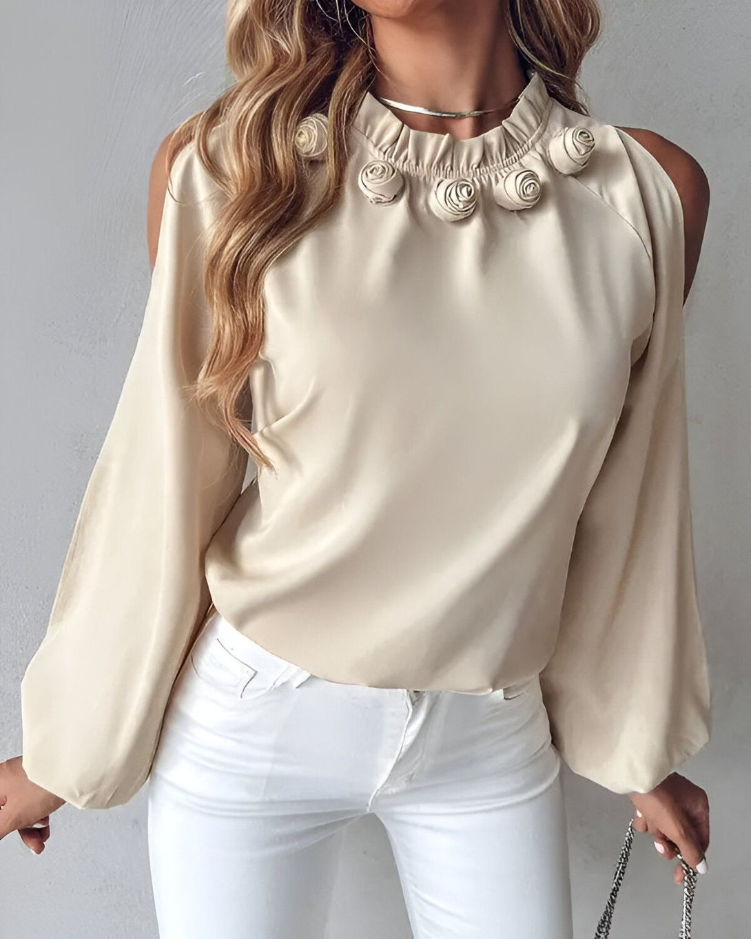 Une femme porte un chemisier beige à col montant et à manches longues. Le chemisier est orné de roses en tissu sur le col. La femme a les cheveux longs et blonds et porte un jean blanc.