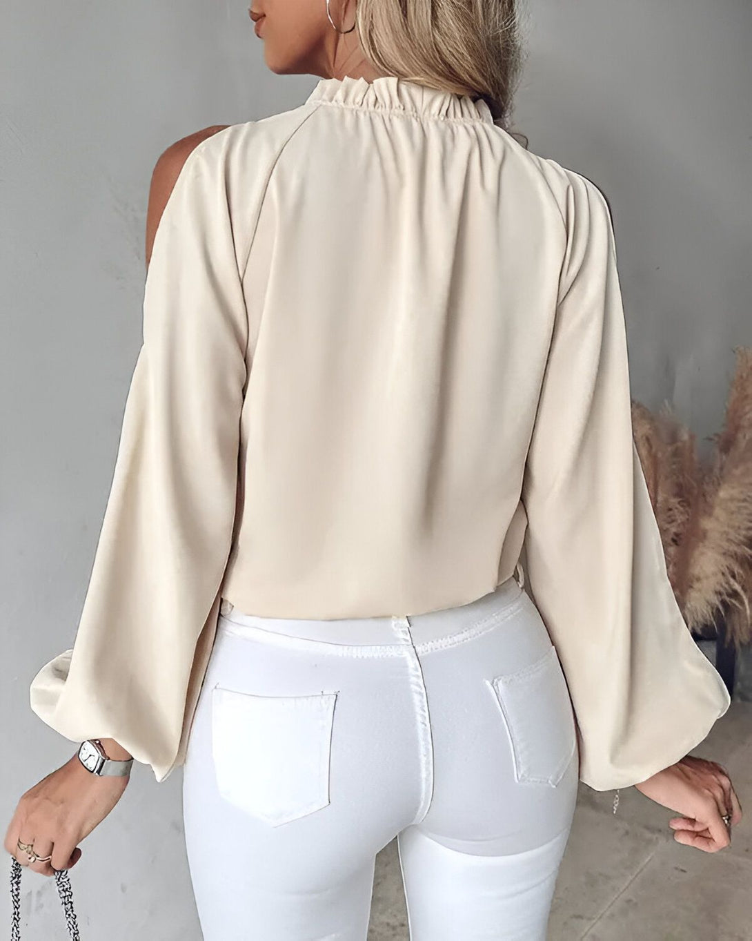 Une femme porte un chemisier beige à une épaule et un jean blanc. Le chemisier est ample et a des manches bouffantes. Le jean est moulant et a une taille haute. La femme est blonde et a les cheveux longs. Elle porte un collier et un bracelet. Elle est debout et regarde par-dessus son épaule droite.