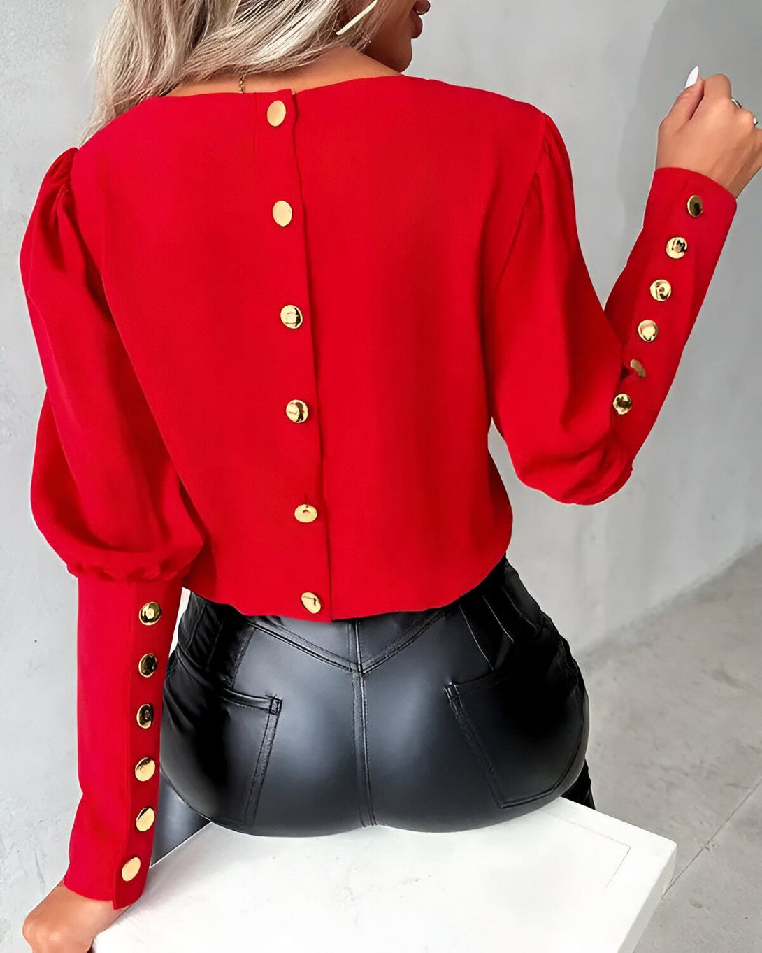 Une femme porte un chemisier rouge avec des boutons dorés dans le dos. Le chemisier est court et les manches sont bouffantes. La femme porte également un pantalon en cuir noir.