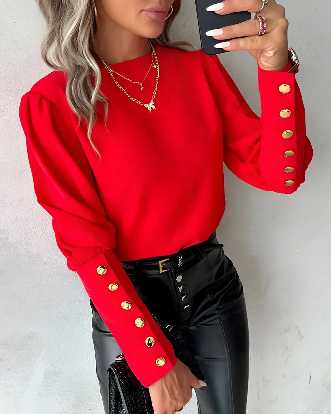 Une femme porte un chemisier rouge à manches bouffantes et boutons dorés. Elle porte également un pantalon en cuir noir et un collier avec un pendentif papillon.