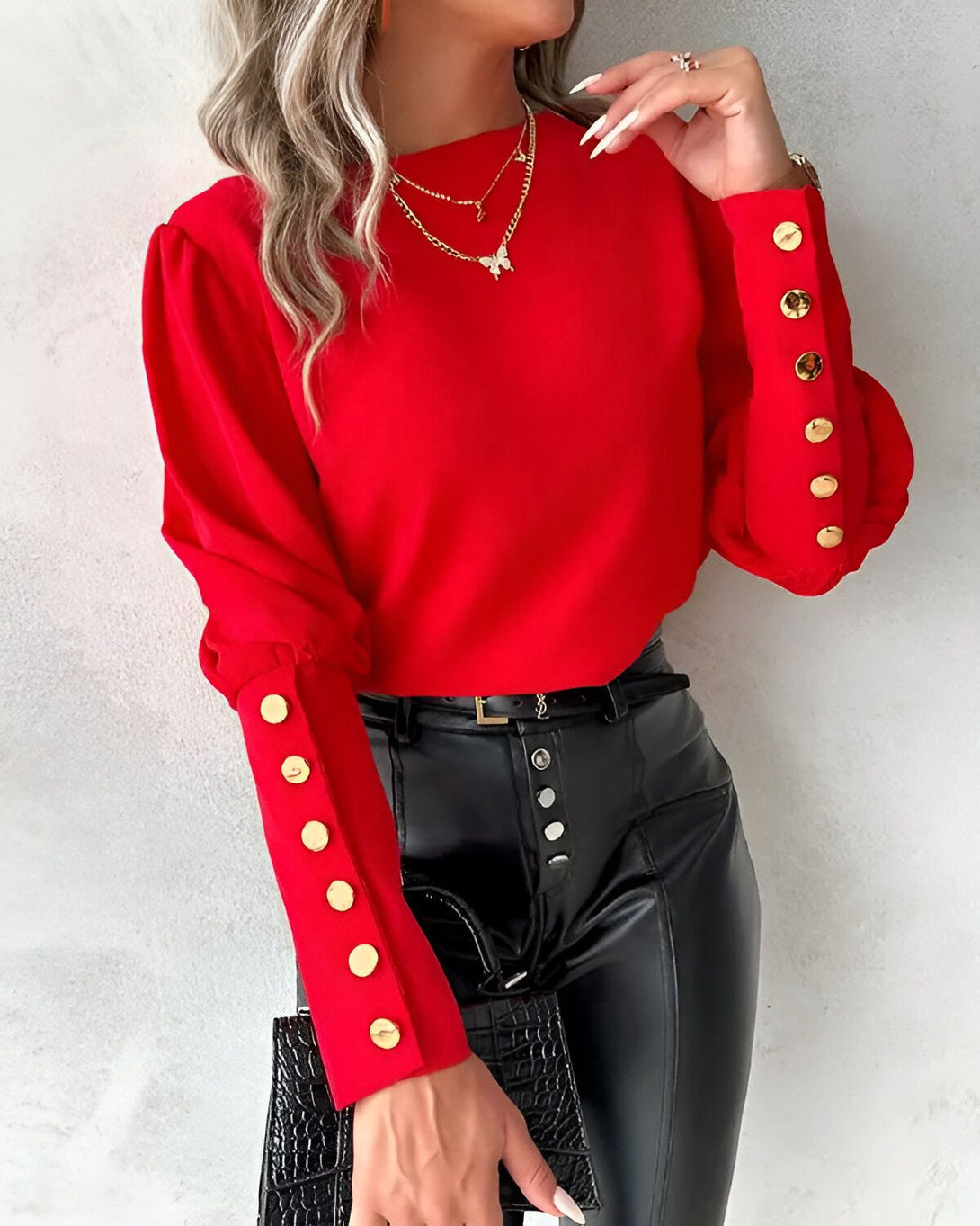 Une femme porte un chemisier rouge à manches bouffantes et des boutons dorés. Elle porte également un pantalon en cuir noir et un collier avec un pendentif papillon.