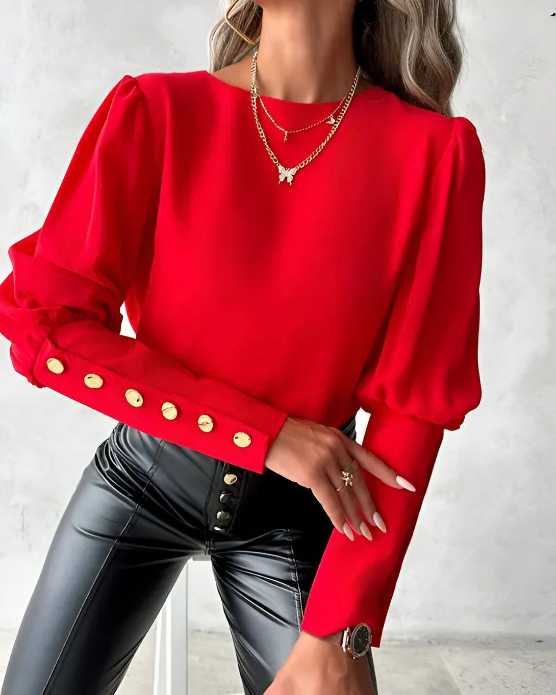 Une femme porte un chemisier rouge à manches bouffantes et des boutons dorés. Elle porte également un pantalon en cuir noir et un collier avec un pendentif papillon.