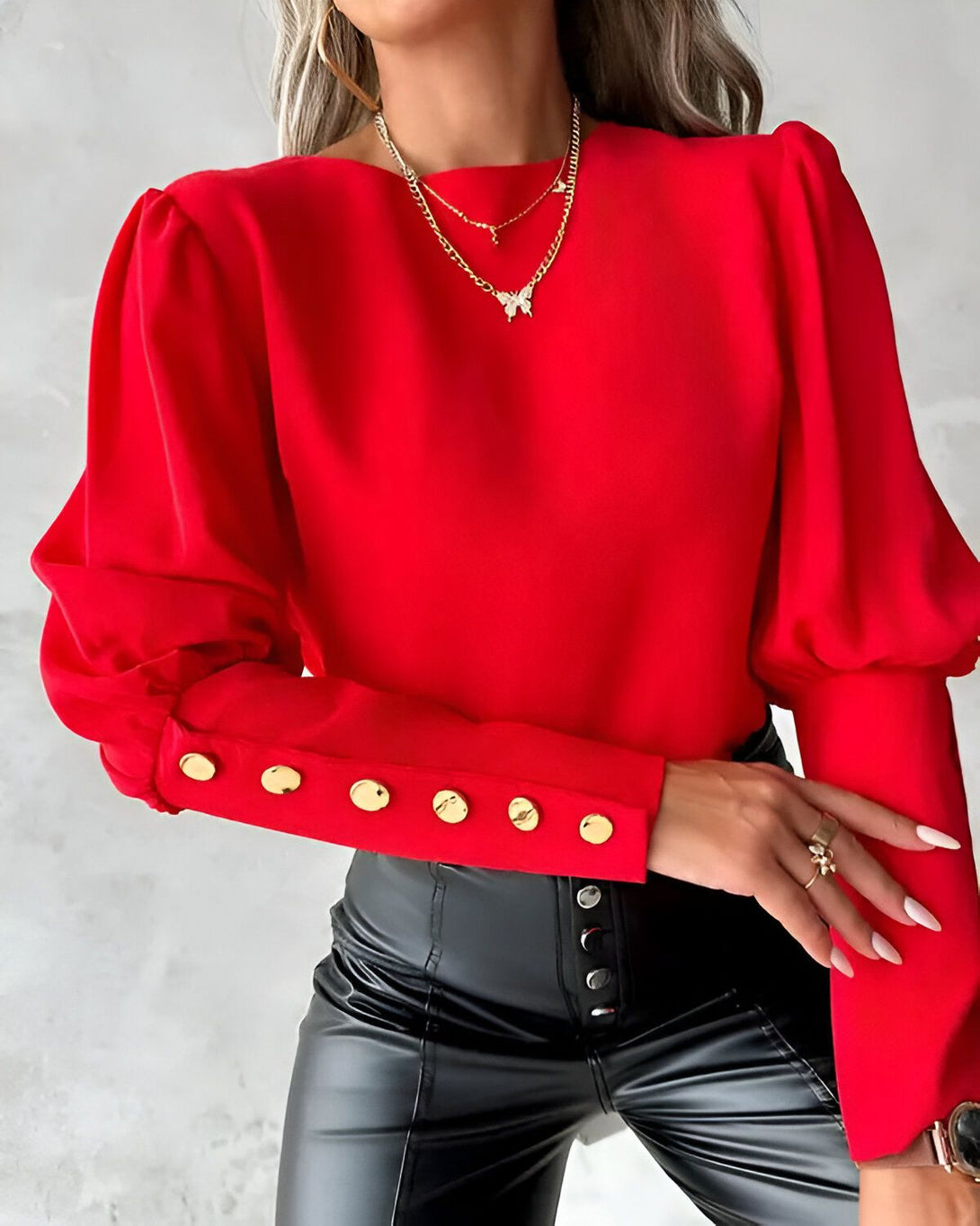 Une femme porte un chemisier rouge à manches bouffantes et des boutons dorés. Elle porte également un pantalon en cuir noir et un collier avec un pendentif papillon.