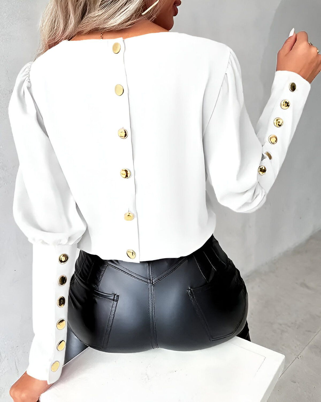 Une femme porte un chemisier blanc avec des boutons dorés dans le dos. Le chemisier est court et les manches sont bouffantes. La femme porte également un pantalon en cuir noir.
