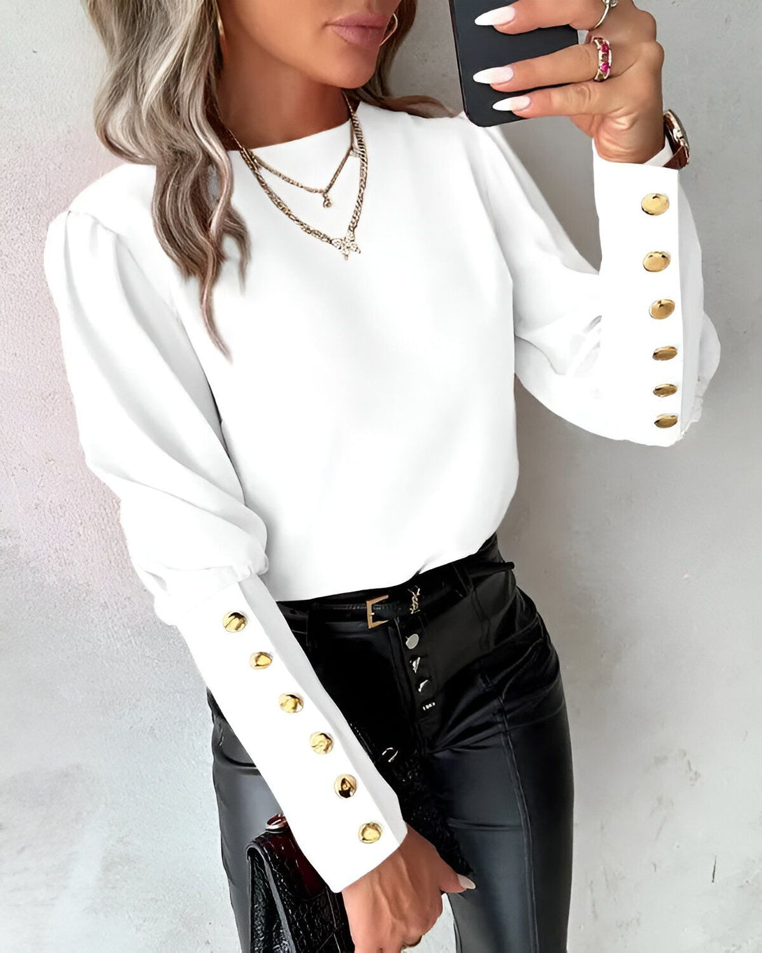 Une femme porte un chemisier blanc avec des boutons dorés sur les manches. Elle porte également un pantalon noir et un collier.