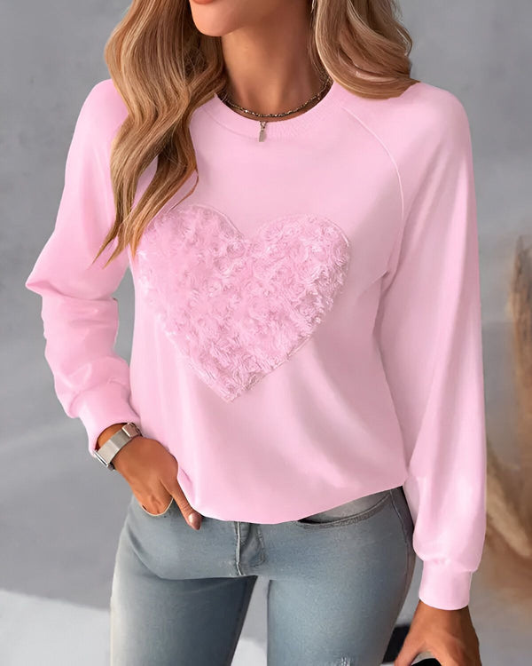 Une jeune femme porte un sweat-shirt rose avec un cœur en fourrure rose sur le devant. Elle a les cheveux longs et blonds et porte un jean bleu.