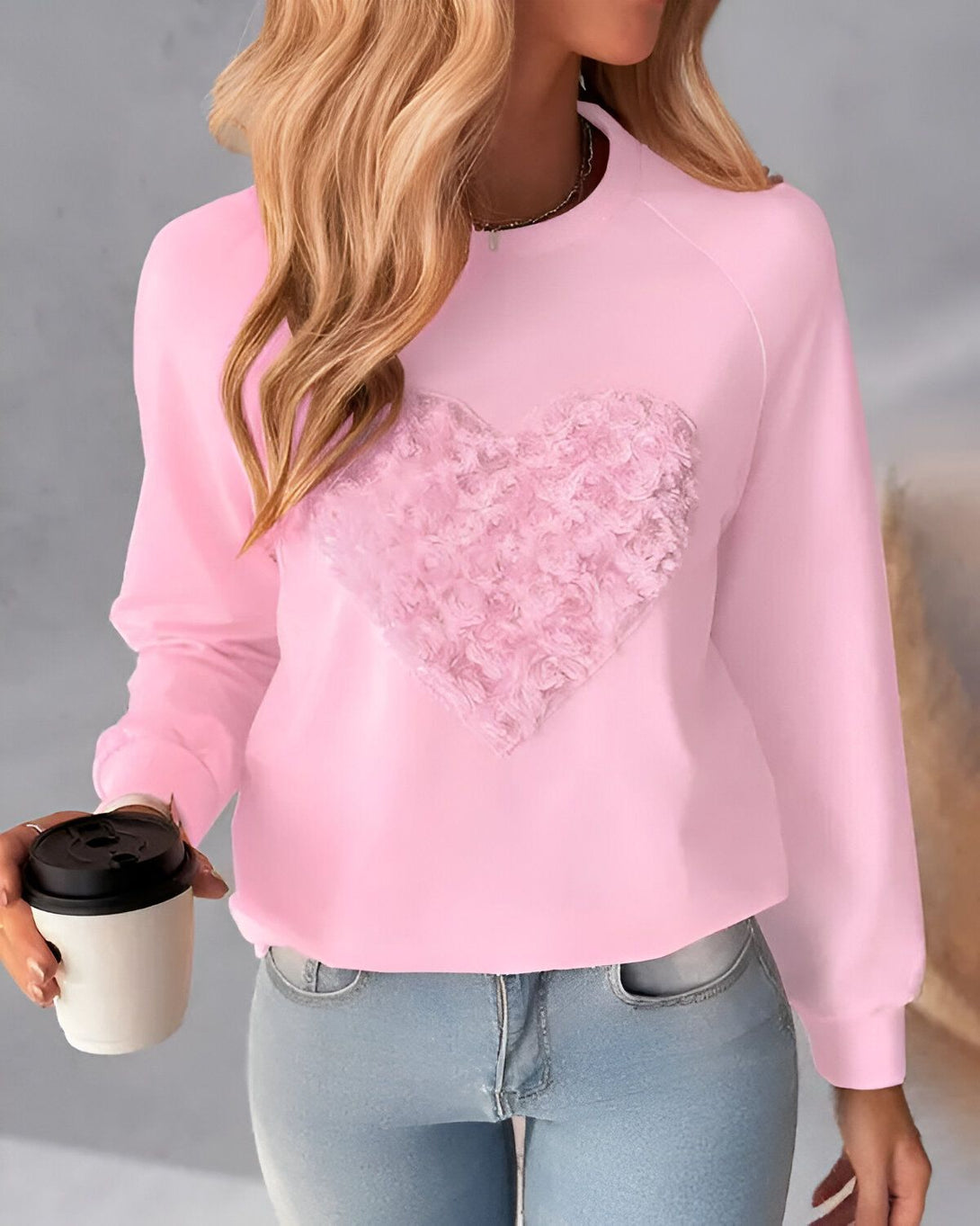 Une femme porte un sweat-shirt rose avec un cœur en relief sur le devant. Elle tient une tasse de café à la main.