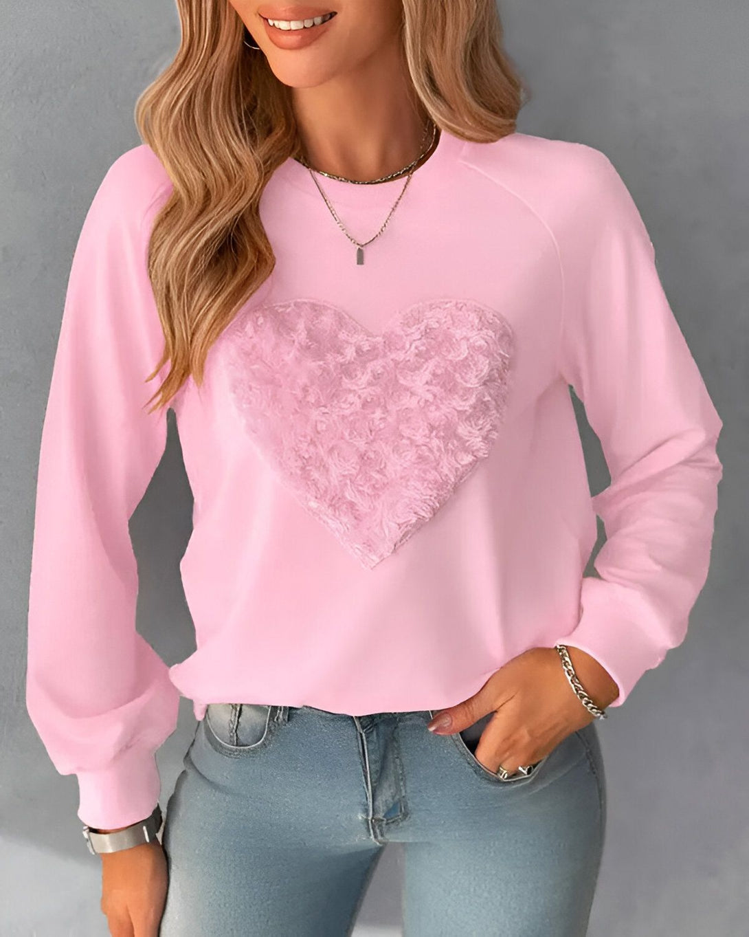 Une jeune femme porte un sweat-shirt rose avec un cœur en fourrure rose sur le devant. Elle a les cheveux longs et blonds et porte un jean bleu.