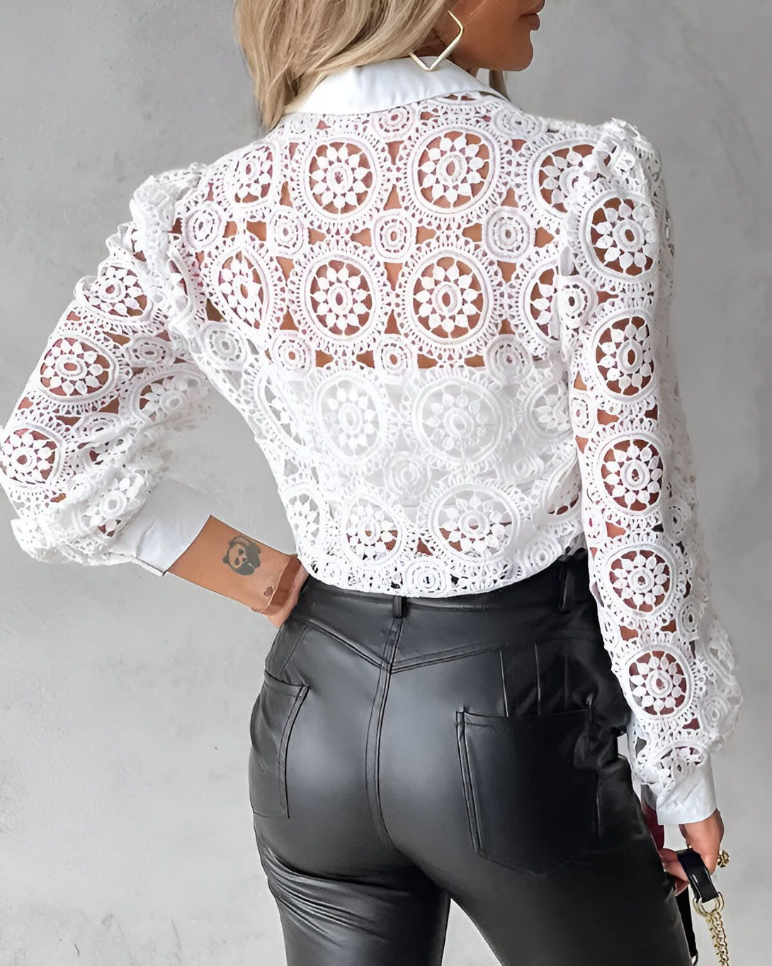 Une femme porte un chemisier blanc en dentelle avec des manches bouffantes et un col montant. Elle porte également un pantalon en cuir noir et des talons hauts noirs.