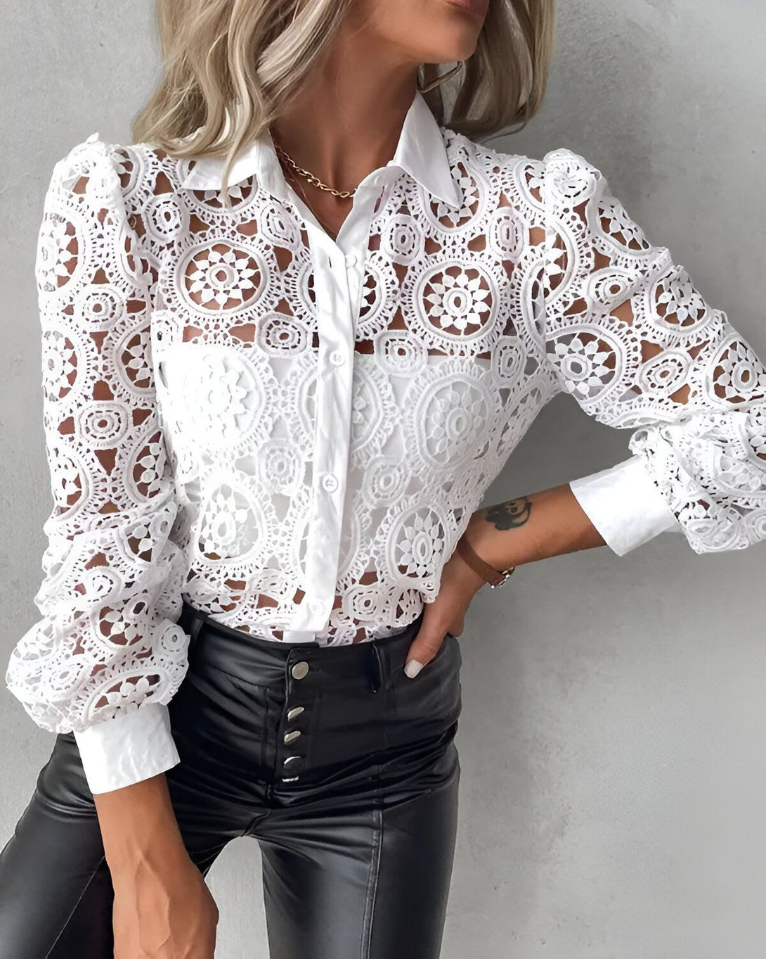 Une femme porte un chemisier blanc en dentelle avec des manches bouffantes et un col montant. Elle porte également un pantalon en cuir noir et un collier.