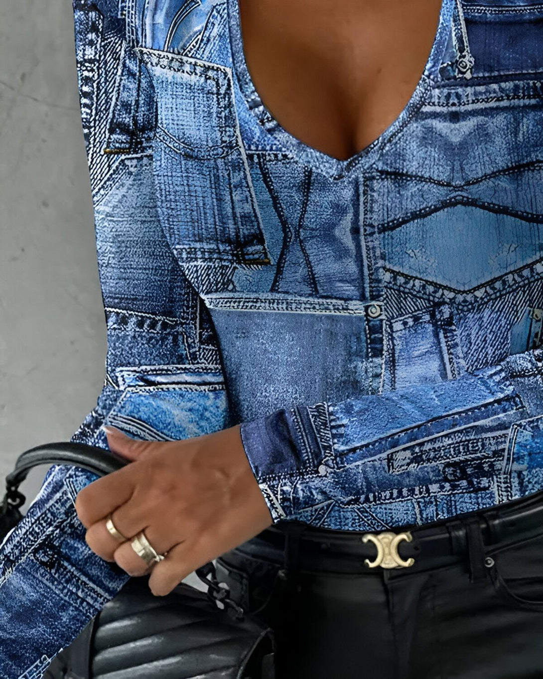 Une femme porte un chemisier bleu à col en V et des manches longues. Le chemisier est imprimé avec un motif de jeans. La femme porte également un pantalon noir et une ceinture noire. Elle a les cheveux longs et noirs et porte du maquillage.