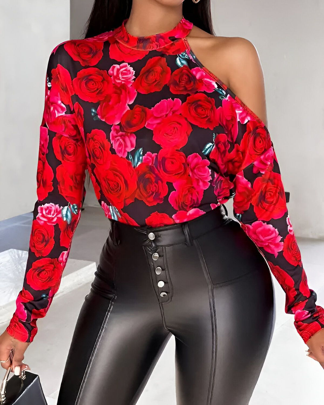 Une femme porte un chemisier noir à imprimé floral rouge et un pantalon en cuir noir. Elle a une épaule dénudée et porte un sac à main noir.