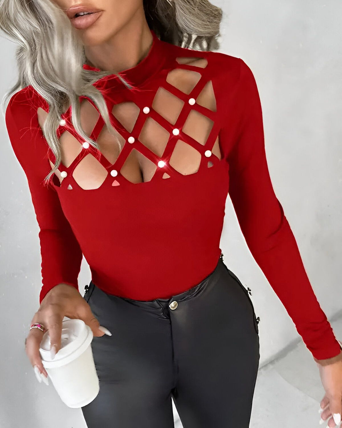 Une femme porte un haut rouge moulant à manches longues. Le haut est échancré au niveau de la poitrine et du ventre, et est décoré de perles. Elle porte un pantalon en cuir noir et tient une tasse blanche à la main.