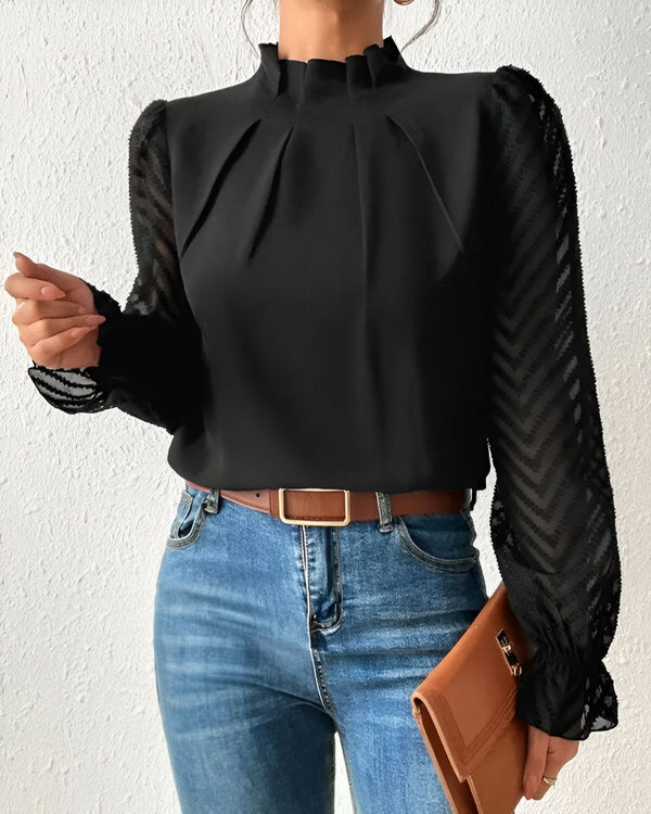 Une femme porte un chemisier noir à manches longues et un jean bleu. Le chemisier est doté d'un col montant et de poignets froncés. La femme porte également une ceinture marron et un sac à main marron.