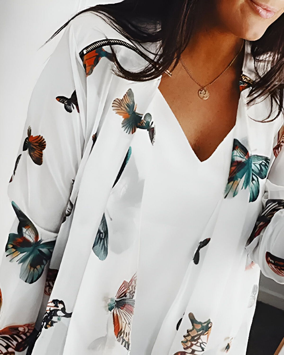 Une femme porte une blouse blanche à motifs papillons. La blouse est ouverte et on voit un t-shirt blanc en dessous. La femme porte également un collier doré.