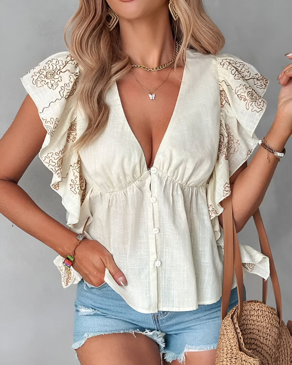 Une jeune femme porte un chemisier ample beige avec des boutons sur le devant et des manches courtes à volants brodés. Elle porte également un short en jean bleu clair et un sac à main en paille marron.
