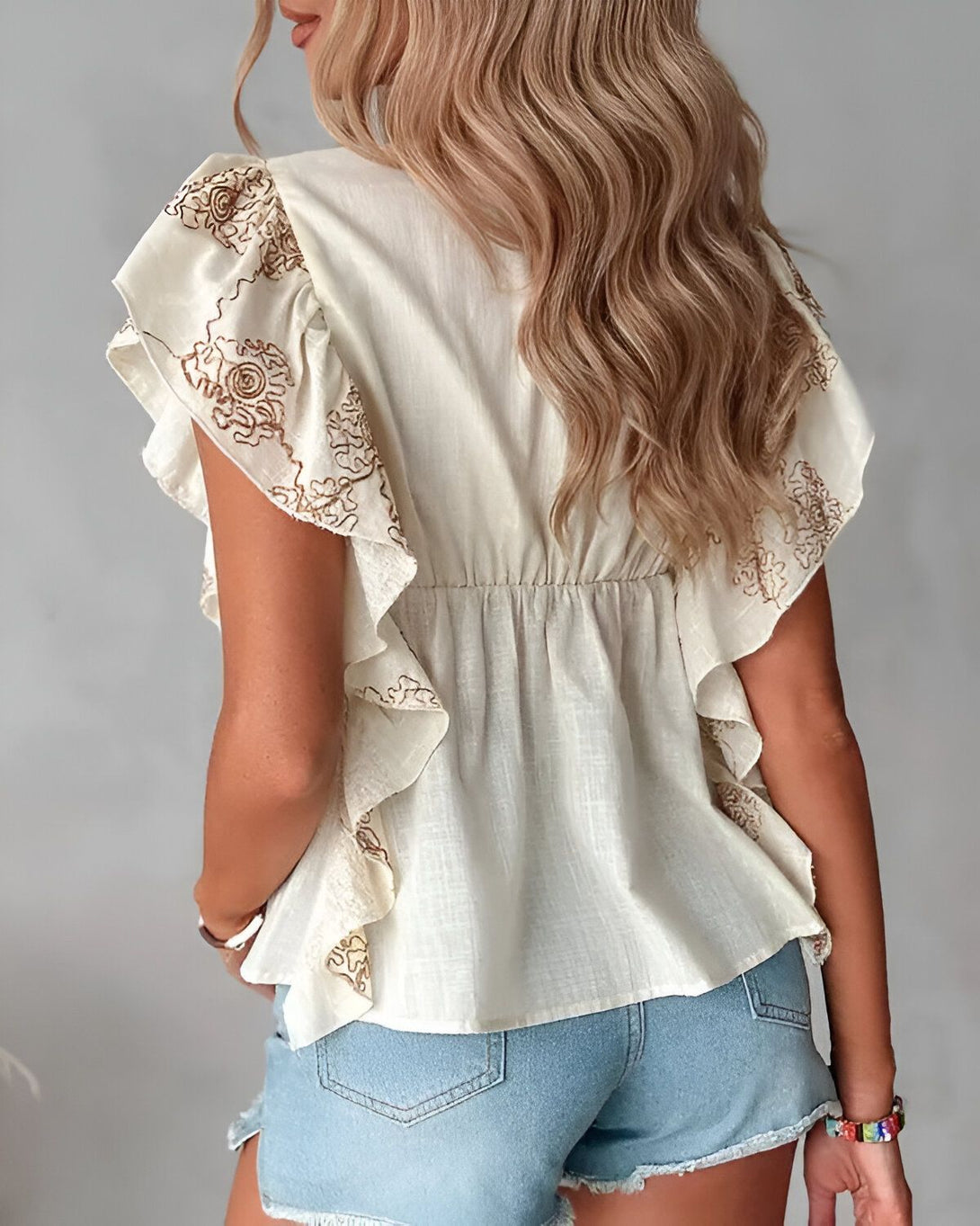 Une femme porte un chemisier ample en coton de couleur beige avec des motifs brodés marron. Il a des manches courtes à volants et un col en V. Elle porte également un short en jean bleu clair taille haute.