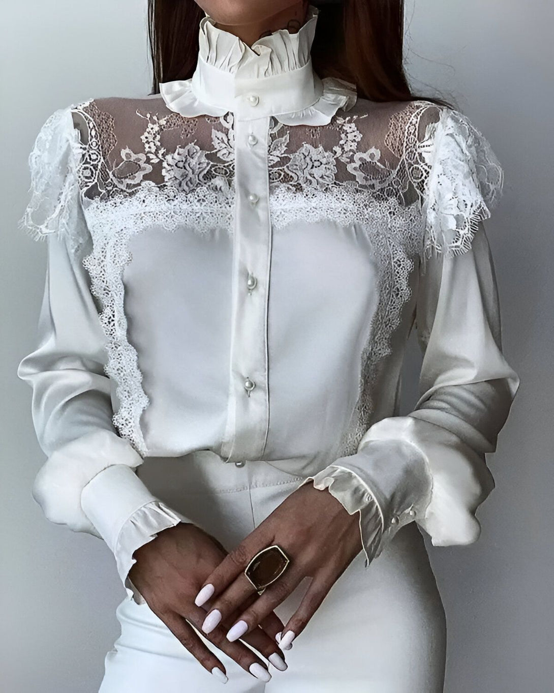 Une femme porte un chemisier blanc en dentelle. Le chemisier est transparent au niveau des épaules et du haut du dos. Il est boutonné sur le devant et possède un col montant. La femme porte également un pantalon blanc.