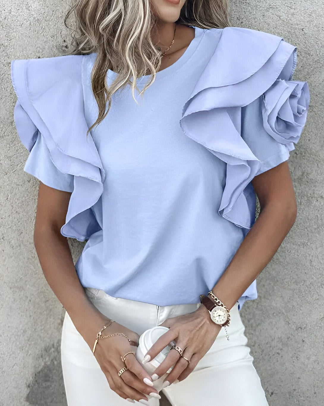 Une jeune femme porte un chemisier bleu à volants. Le chemisier est à manches courtes et possède un col rond. La femme porte également un pantalon blanc et des talons hauts. Elle a les cheveux longs et blonds et porte un maquillage léger.