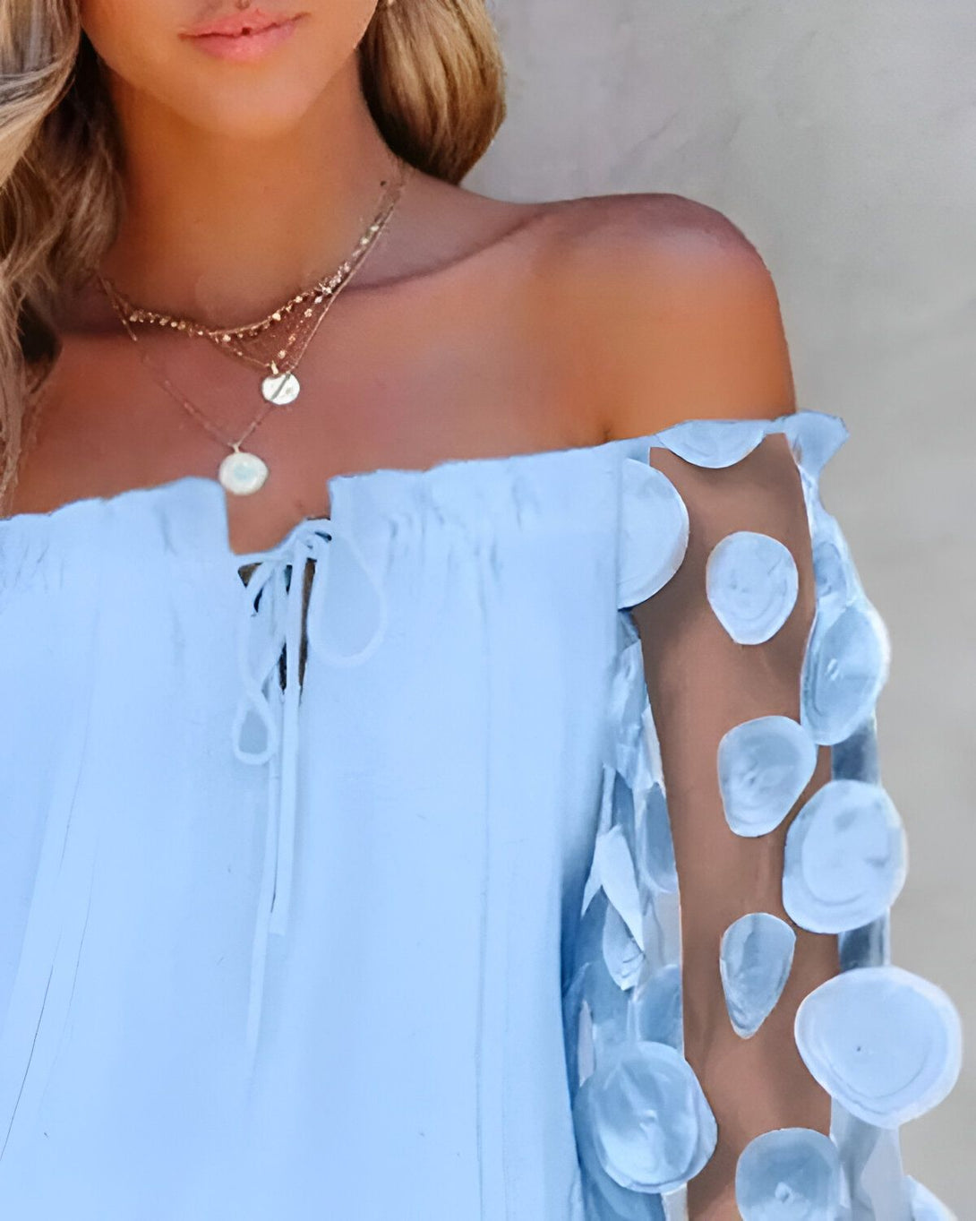 Une jeune femme porte une robe bleu clair à épaules dénudées. La robe est en mousseline de soie et comporte des manches bouffantes. La femme a les cheveux blonds et porte un collier.