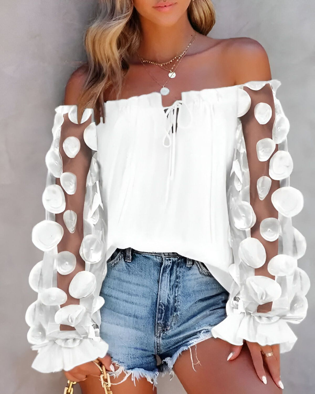 Une jeune femme porte un chemisier blanc à épaules dénudées. Le chemisier est orné de pois blancs en relief. Elle porte également un short en jean bleu et un collier avec un pendentif rond.